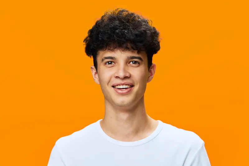 Smiling young man with curly hair wearing a white t-shirt stands against vibrant orange background, showing cheerful mood and casual style in bright studio lighting.