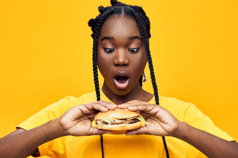 Surprised young Black woman holding a burger with excitement, colorful yellow background brings a vibrant feel to the image, perfect for food related themes
