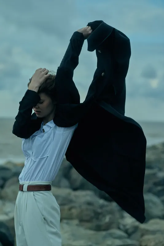 Wind blows cape over a fashion forward silhouette on a rocky shore, a model in white shirt and belt posing dramatically with flowing outerwear under a moody sky