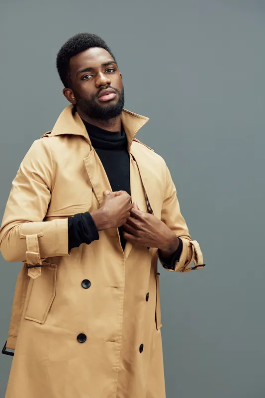 Stylish young man in a beige trench coat posing confidently against a grey background, showcasing modern fashion and seasonal style