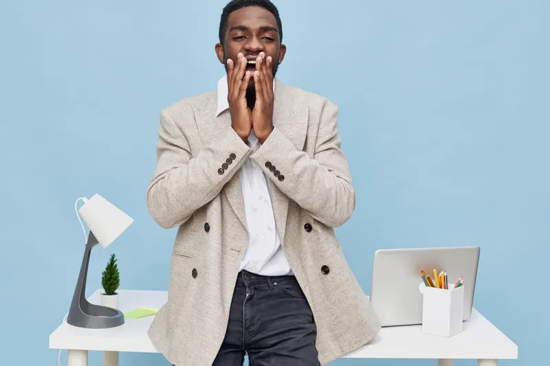 Smiling cheerful man in beige blazer and white shirt standing at desk with laptop and stationery on blue background. People lifestyle concept, happiness, success, workspace, positive emotions, adult m