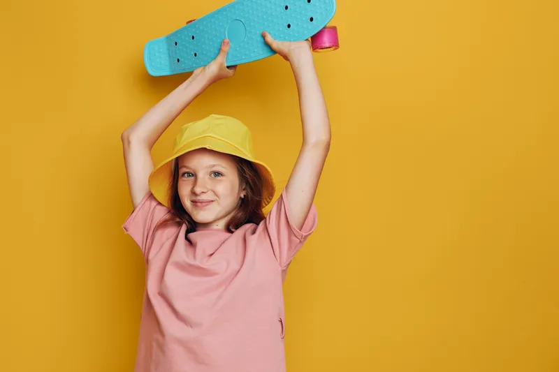 Smiling girl in yellow hat holding blue skateboard above head on orange background. Happy child with casual pink t shirt studio portrait.