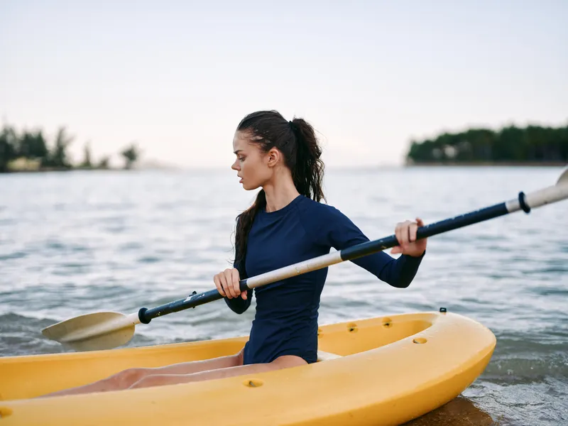 Female kayaker sits in a yellow kayak, gripping a paddle as she glides across calm water, with distant shoreline and trees on the horizon.