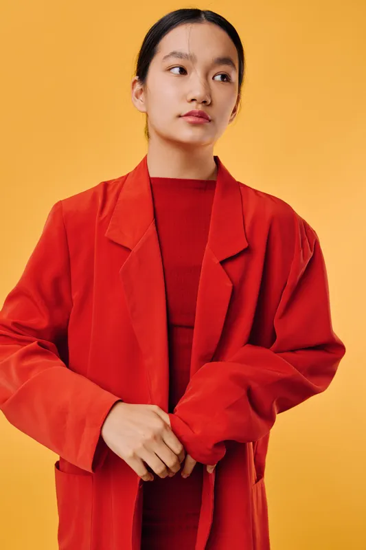 Woman with black hair wearing oversized red coat and red dress standing against orange background, looking sideways with folded hands in casual fashion portrait with minimalist style.