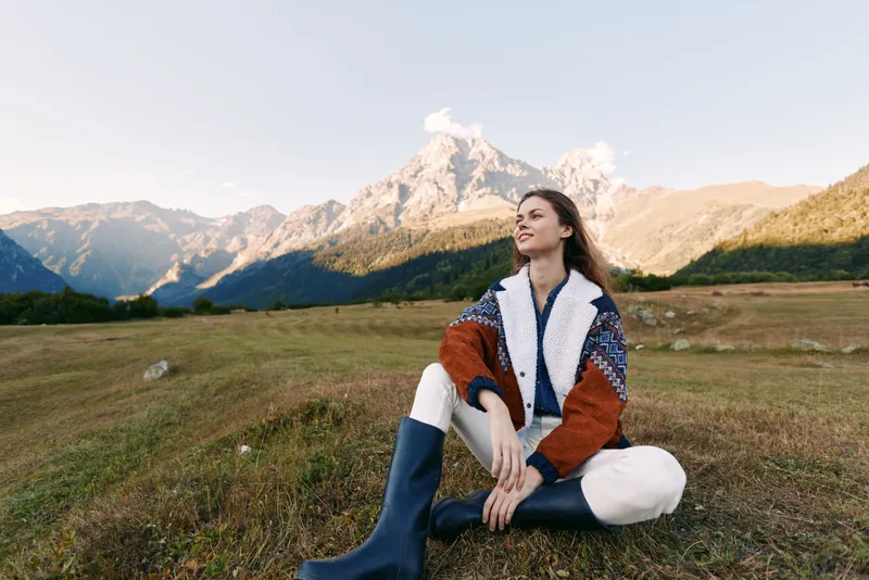 Woman in autumn fashion sitting on a meadow outdoors with mountains in background, wearing boots and warm jacket, relaxed portrait of travel, nature and scenic landscape.
