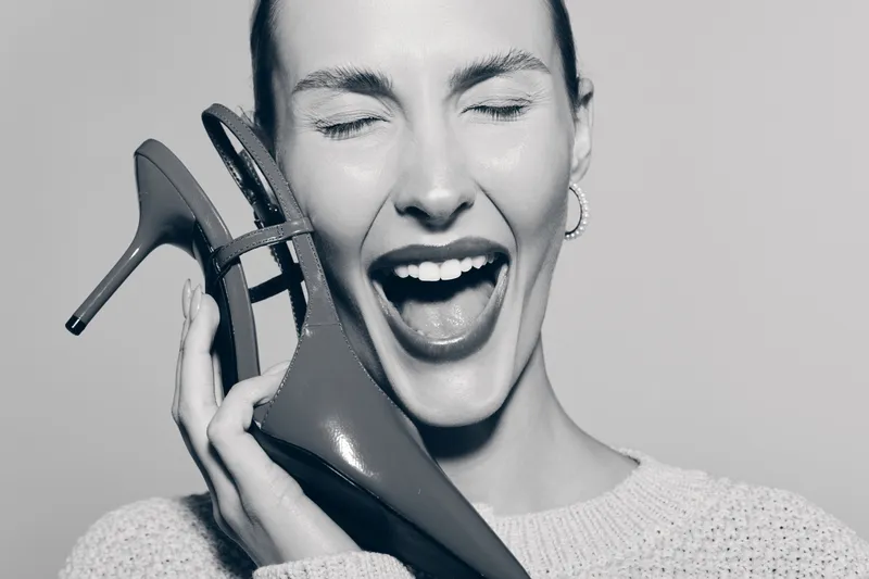 Happy Caucasian woman with light skin holding high heel shoe near face smiling with closed eyes in black and white. Concept of fashion and joy.