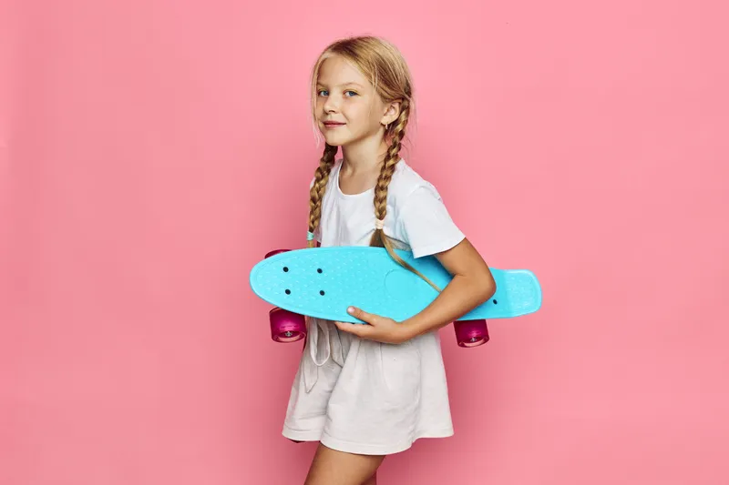 Smiling young girl with blonde hair in braids holds a bright blue skateboard. She wears casual white and beige clothes against a pink background. Concept of childhood and fun.