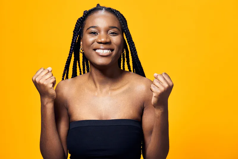 Happy young Black woman with braids expressing joy against a bright yellow background, showcasing vibrant energy and positivity in a fashion forward outfit