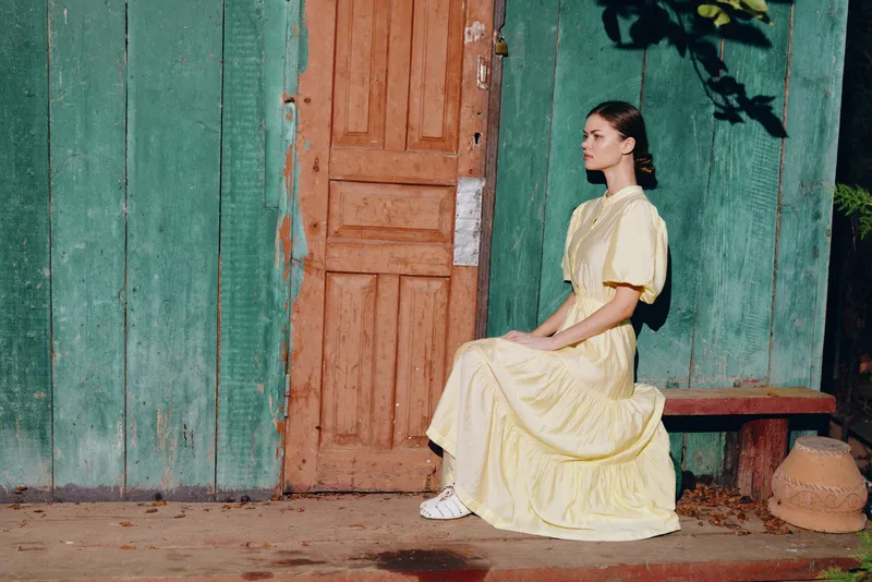 A woman in a pale yellow dress sits on a wooden bench beside a weathered doorway and turquoise wall, capturing a calm rustic moment with soft light