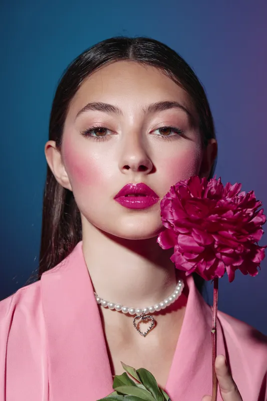 Young woman with bold pink lipstick and blush holds a bright pink flower, contrasting with her pale pink jacket and delicate pearl necklace. Studio fashion beauty portrait.