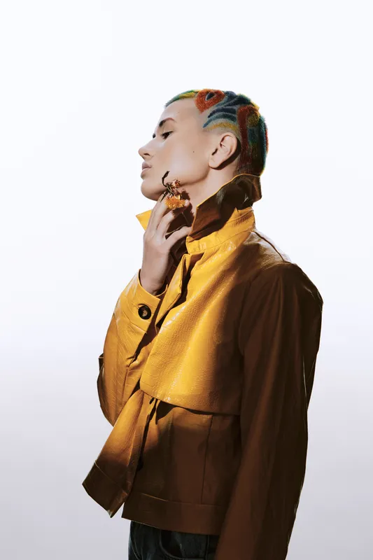 Stylish woman with colorful braided hair and yellow jacket poses thoughtfully in profile against white background, showcasing modern fashion and bold accessories.
