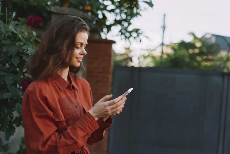 Cheerful woman outdoors, rust shirt, checking phone in sunlit garden, relaxed lifestyle moment, natural colors, greenery background