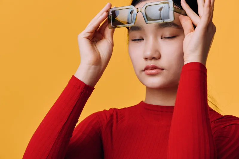 Young woman with closed eyes wearing stylish sunglasses and red sweater posing on yellow background for fashion portrait and casual attitude concept.