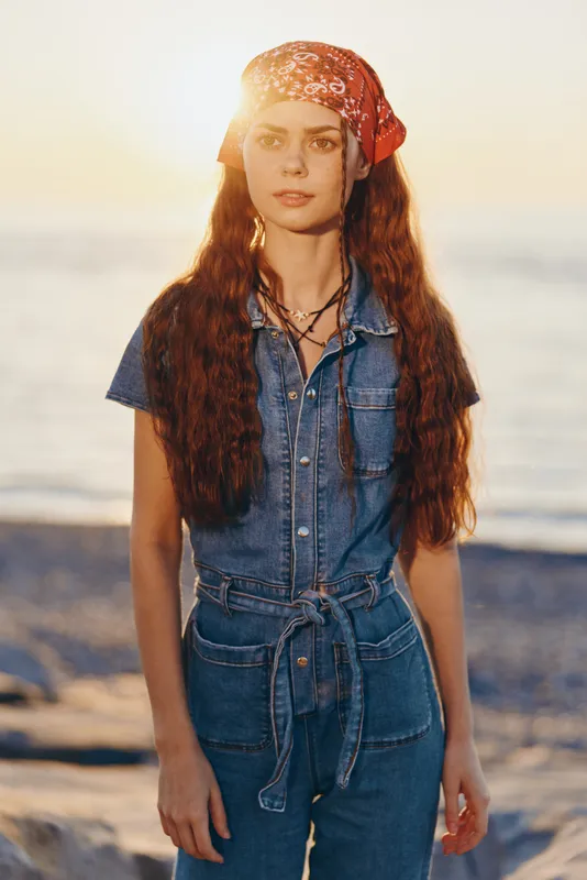 young woman standing on beach at sunset wearing denim jumpsuit and red bandana with long wavy hair backlit by warm sunlight
