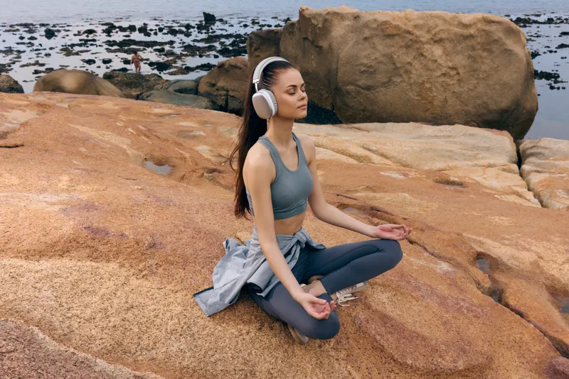 Young woman meditating peacefully on rocky beach with headphones, embodying tranquility, beauty, and wellness in an outdoor setting