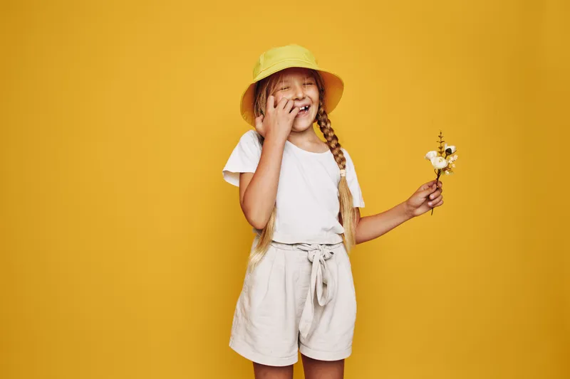 Happy girl in a yellow hat smiling and holding small flowers against a yellow background. She wears white shirt and beige shorts with tied belt. Bright cheerful studio portrait.