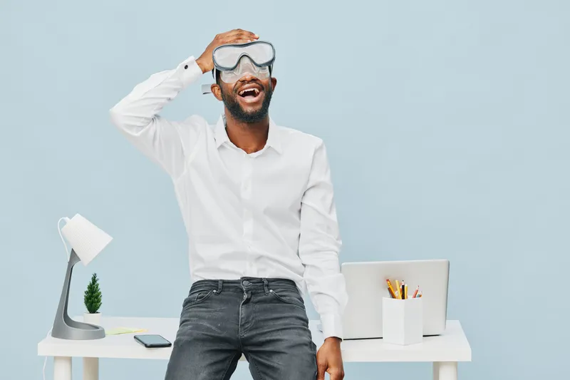 Smiling man with beard wearing protective goggles and white shirt standing in modern office with laptop and desk, enjoying work or fun moment, full body, casual style, bright background, office lifest