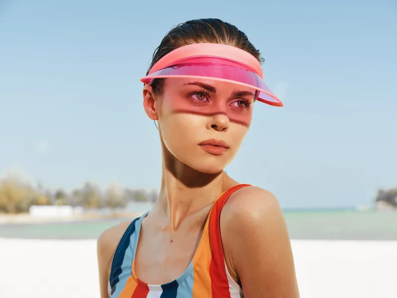 Portrait of a young woman wearing a pink visor and colorful striped swimsuit under bright sunlight, capturing athletic style, focus, and confident gaze against a clear blue sky and distant shoreline,