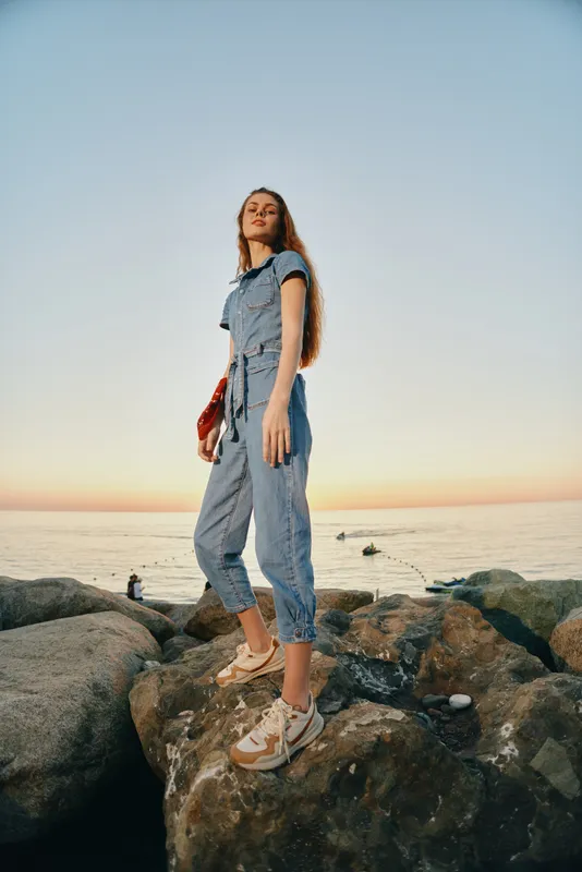 Young woman standing confidently on rocky shore during sunset wearing casual jumpsuit and sneakers near calm sea background, creative outdoor summer scene
