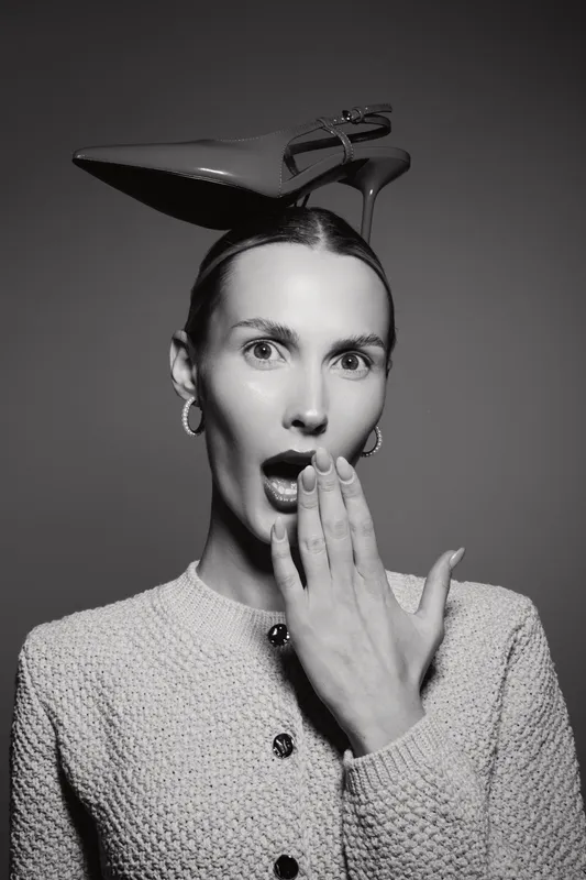 Surprised young caucasian woman with fair skin and dark hair holds hand on mouth with shoe on head.