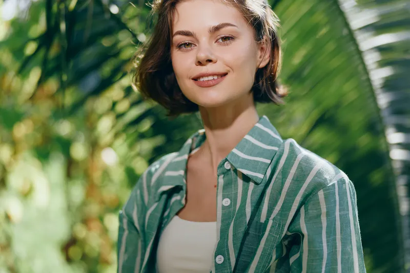 A young woman smiles softly outdoors, wearing a striped green shirt under warm sunlight with lush palm leaves in the background.