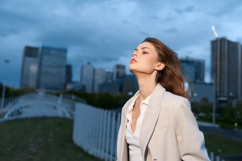 confident woman in beige coat outdoors at dusk with urban buildings, thoughtful expression, autumn fashion, evening cityscape background