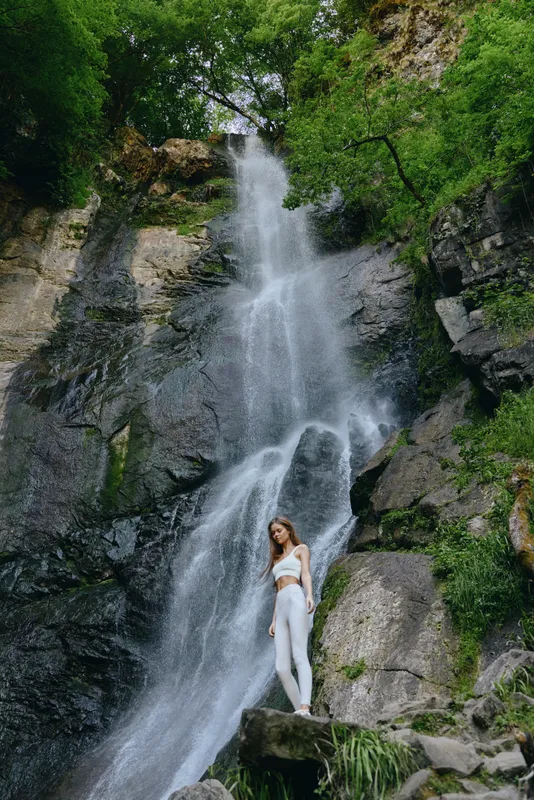 Woman standing near tall waterfall surrounded by lush green forest and rocky cliffs, wearing white activewear, showcasing nature and outdoor adventure.