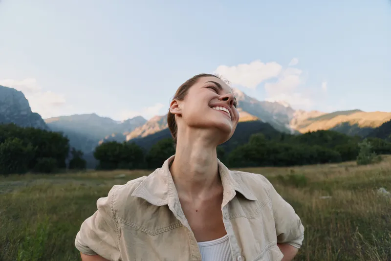 A happy woman in casual clothes stands in a field with mountains and trees in the background, smiling and looking up. Concept for lifestyle, nature, and freedom.