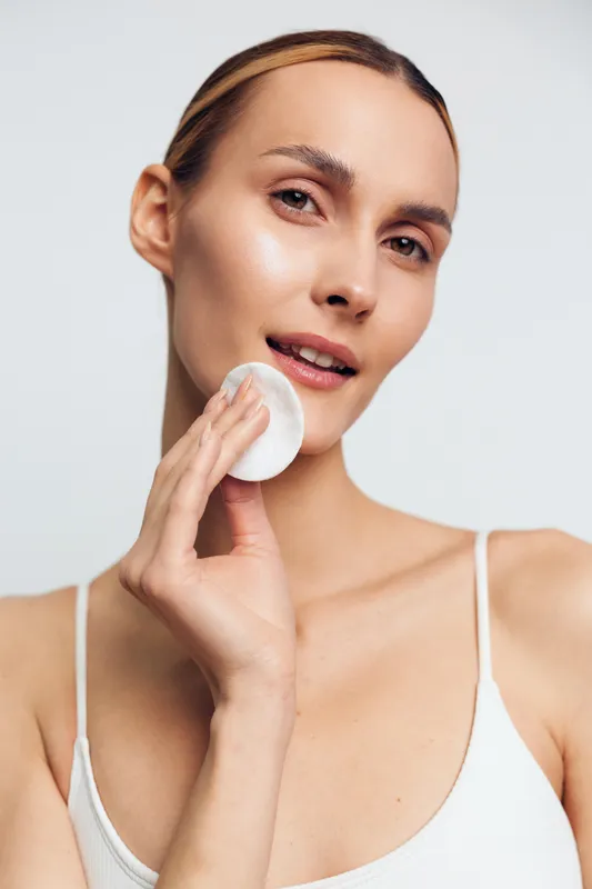 Young woman skincare beauty routine, holding a cotton pad to her face, performing cleansing for hygiene and self care, minimal studio background, calm skin care concept today