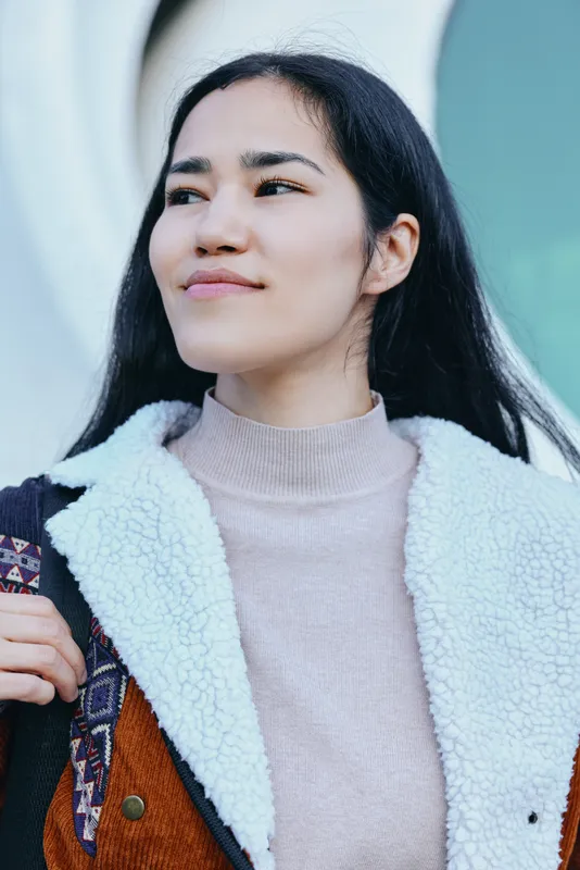Woman wearing a fluffy white-lined coat and colorful jacket, beige sweater, outdoors, presenting a casual fashion mood with a confident smile and street style, soft lighting and urban ambiance