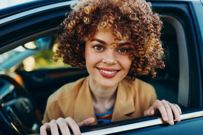 Cheerful woman in car, sunlight on face, relaxed posture, retro film color, lifestyle concept, bright natural tones, candid moment inside vehicle interior and warm inviting mood overall