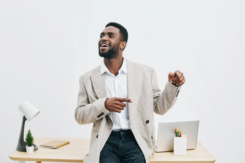 Confident young African-American man in light grey blazer and white shirt pose happily at modern workspace with white wall, desk, laptop, and stationery, bright professional atmosphere, people lifesty
