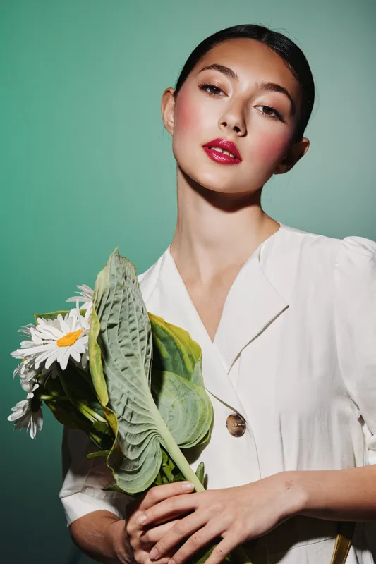 Young woman with red lipstick and glossy skin poses against turquoise background wearing white blouse holding green tropical leaves and daisies. Concept of beauty creativity and fashion studio