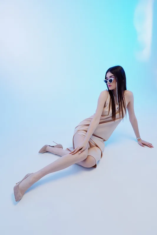 Young woman in beige dress and sunglasses poses elegantly on white floor against blue background. Concept of woman beauty fashion and modern style.