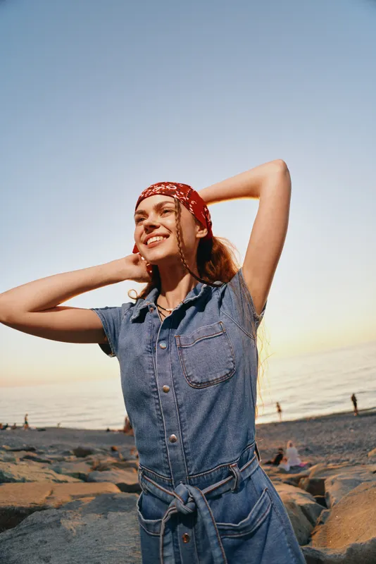 Lifestyle woman in modern boho-western denim street style smiles joyfully outdoors at sunset beach with film color effect, casual and carefree mood captured perfectly.
