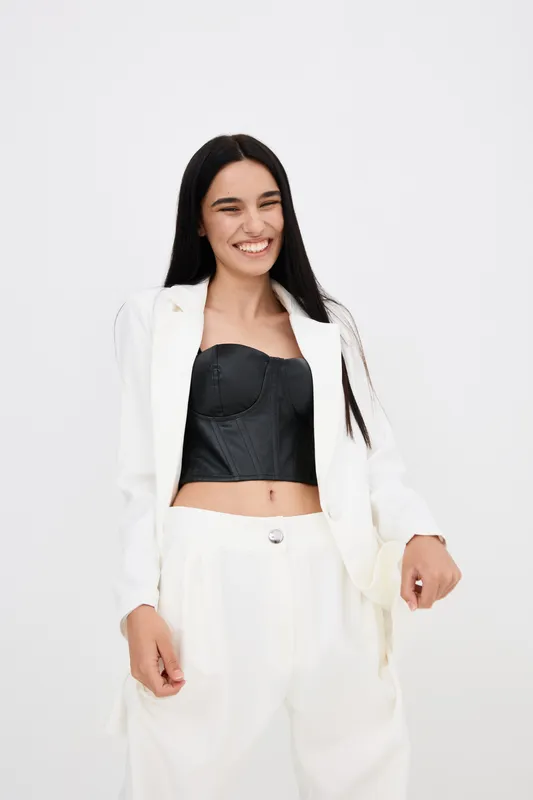 Smiling woman dressed in white suit and black corset top stands against plain light background, showing carefree attitude and joyful expression with long black hair.
