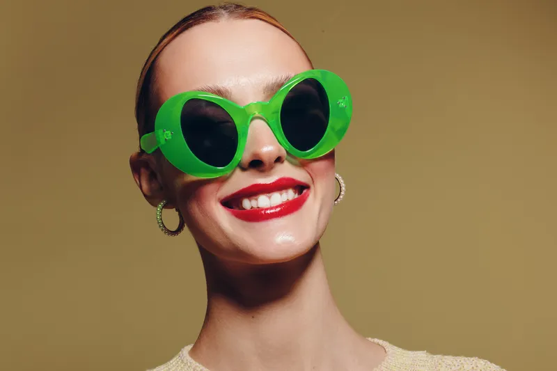 Smiling woman with bright red lipstick wears large green sunglasses and hoop earrings. Close-up portrait on beige background. Concept of fashion, style, and summer accessories.