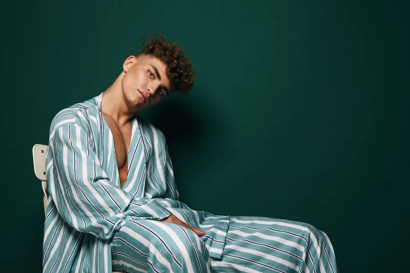 Young man with curly hair wearing striped pajamas sitting on chair against dark green background in studio. Casual relaxed pose with thoughtful expression and soft lighting on face.