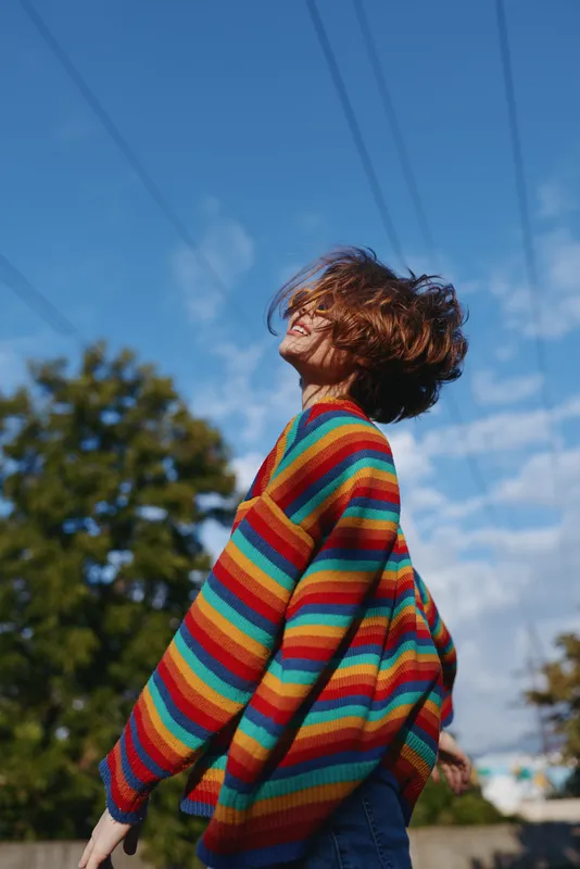 Young woman in a rainbow striped sweater smiling outdoors under a clear blue sky, short hair blowing in the wind, casual outfit and relaxed portrait capturing joyful travel lifestyle energy.