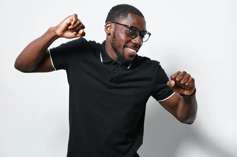 African American man with dark skin wearing glasses and black polo shirt dancing and smiling on white background. Happy young male enjoying music and movement in casual outfit.