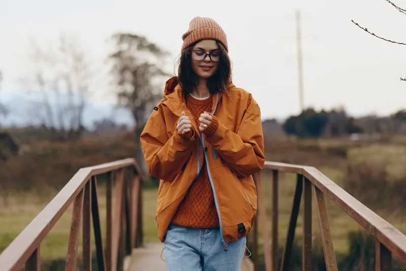 Woman lifestyle portrait outdoors, walking on a wooden bridge in autumn countryside, wearing an orange jacket, knit beanie, scarf and glasses, relaxed smile, warm casual style, blurred nature