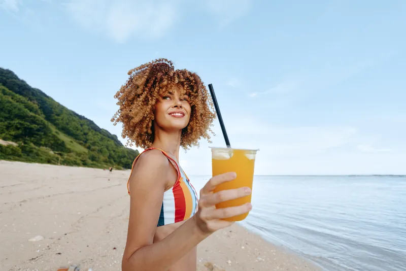 Young woman with curly hair holding a cup of orange juice on a sandy beach by the ocean, sunny summer mood, relaxed smile, coastal landscape with green hills, outdoors warm light and clear sky