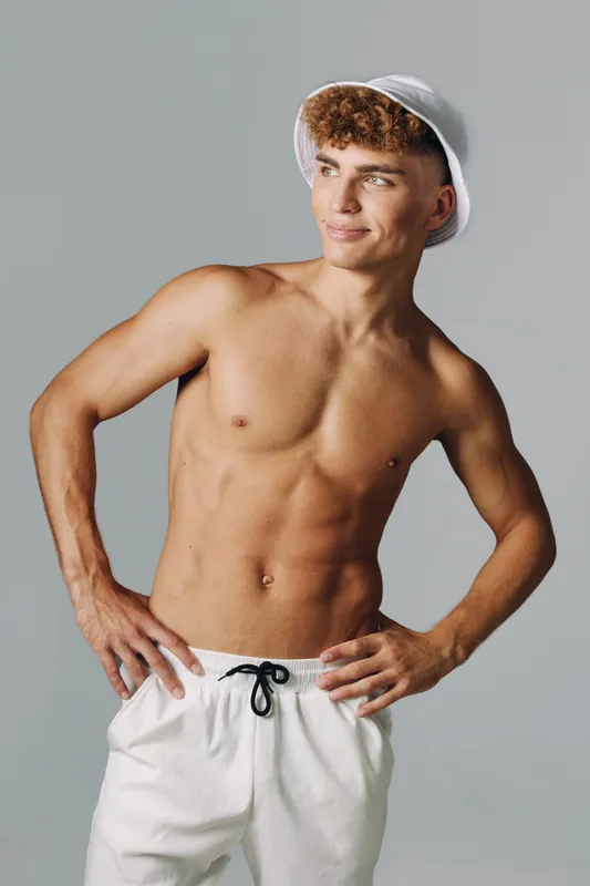 Young man with muscular body posing shirtless wearing white shorts and bucket hat, looking sideways with confident smile on gray background in studio lighting.