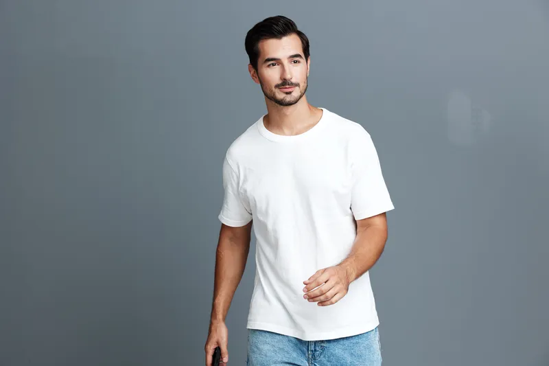 Casual young smiling man wearing a white t-shirt walking confidently against a grey background, holding a black mobile phone in his hand. Portrait lifestyle concept.