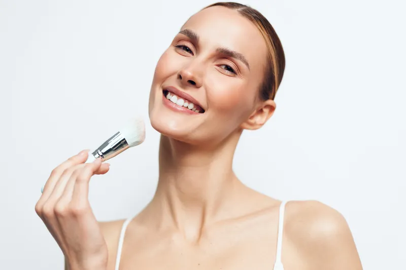 Smiling woman applying makeup with a brush, clear skin, natural light, close-up portrait, beauty routine, white background, clean look, casual style, fresh appearance