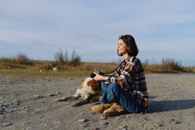 Woman sitting on a sandy beach with a camera, enjoying outdoor leisure with her dog, calm sky and warm natural light in a peaceful coastal moment of connection