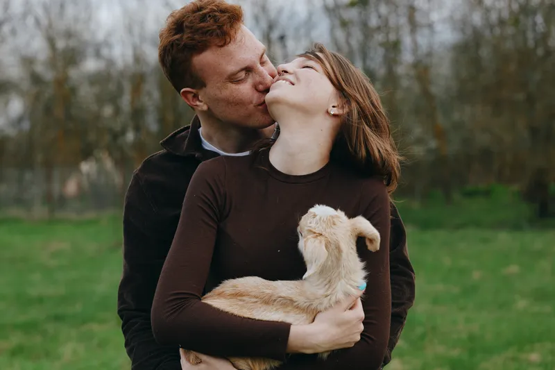 Happy couple shares a tender kiss outdoors, holding a young goat kid in a park setting, showcasing warm affection, candid lifestyle, and romantic charm in natural light.
