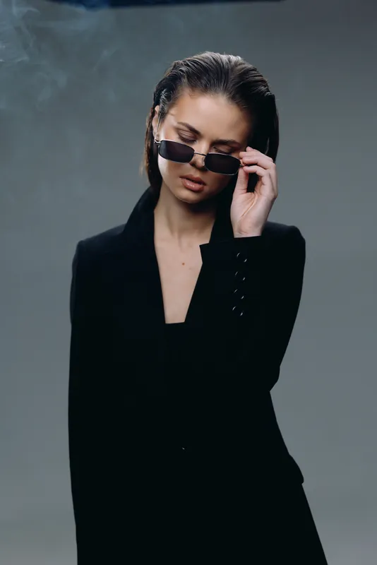 Stylish woman wearing sunglasses and black blazer with serious expression in neutral studio background
