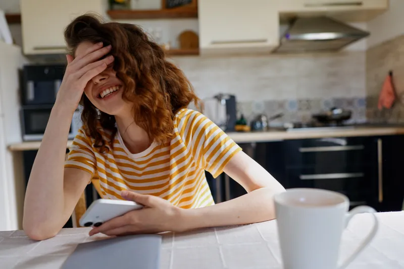 Joyful young woman with curly hair laughs at home while scrolling her phone at a bright kitchen table, wearing a yellow striped t shirt, candid everyday moment.