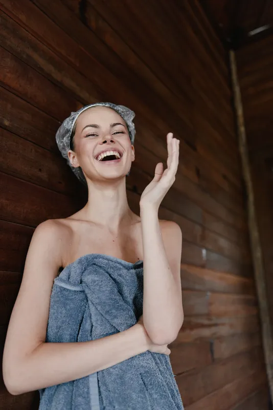 laughing woman wrapped in towel with hair covered standing against wooden wall enjoys spa moment and relaxation, natural skin, wellness concept